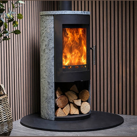 Cylindrical grey soapstone stove with black frame glass door showing fire and integrated log store holding wood
